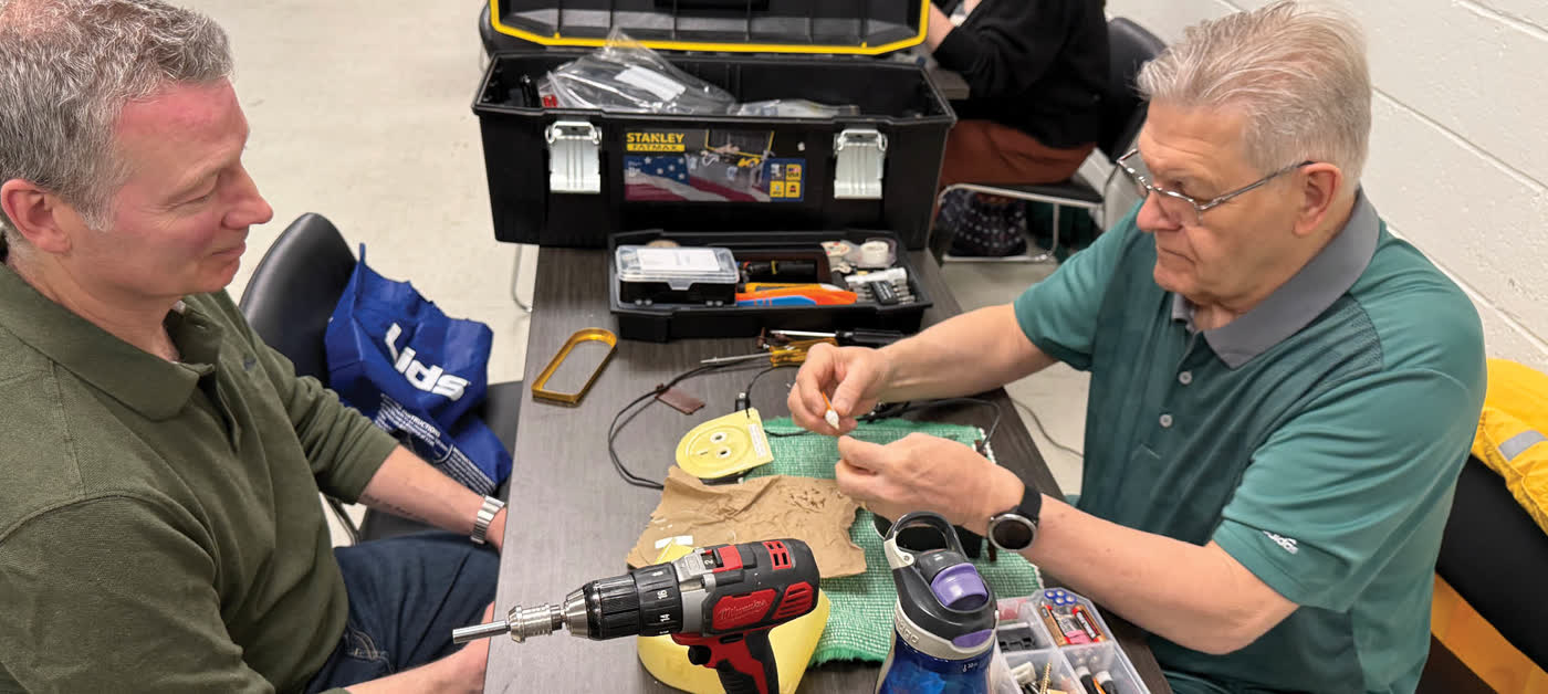 A volunteer fixer works on a small appliance while its owner watches, at a London Repair Café table covered in tools.